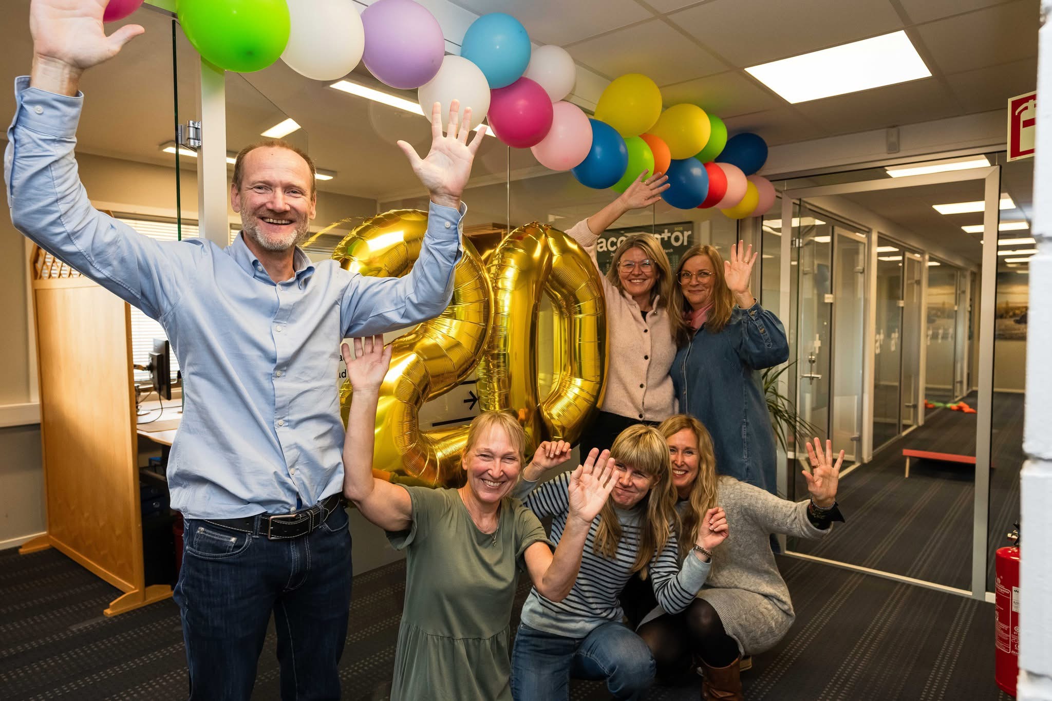 Paul Arne og hans ansatte foran 20 års banneren med fargerike ballonger. Alle har hendene i været og smiler bredt.