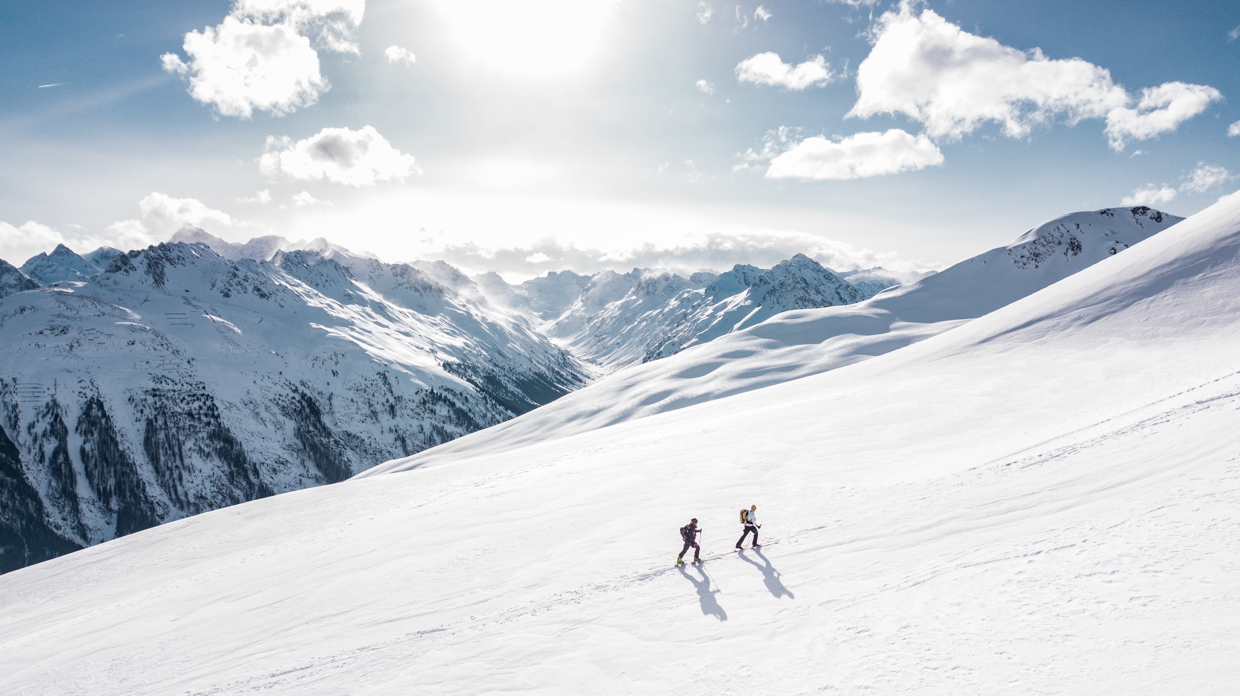 2 mennesker som går på ski oppover fjellet mens solen steker. Fjell i bakgrunnen med litt snø på toppen
