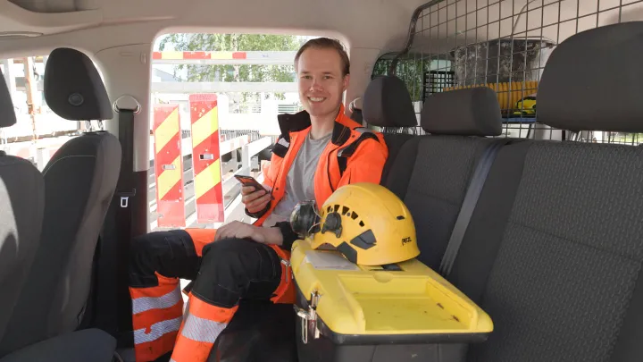 Mann sitter smilende bak i bilen med verneutstyr og holder på med telefonen