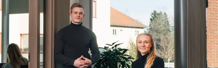 Ung mann og kvinne som står og smiler mot kamera i et kontorlandskap. hun sitter til høyre for han i en stol, mens hans står til venstre. Begge har på seg en svart turtleneck og blå jeans. 