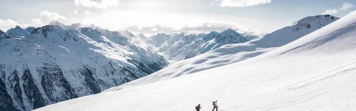 2 mennesker som går på ski oppover fjellet mens solen steker. Fjell i bakgrunnen med litt snø på toppen 