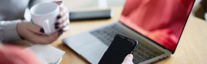 To kvinner som sitter sammen over en laptop. den ene holder i en telefon. Man kan kun se armene deres ingen ansikter.